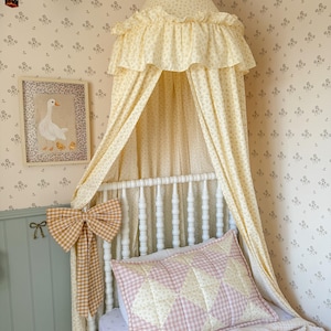 May include: A yellow floral fabric bed canopy with a ruffled top, suspended over a white spindle bed frame. A quilted pillow with a pink and yellow diamond gingham pattern rests on the bed, accented by a large yellow and white gingham bow.