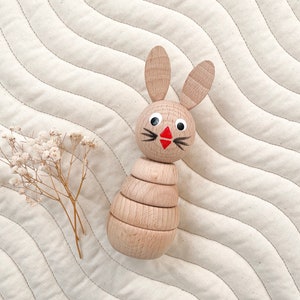 May include: A wooden stacking toy shaped like a bunny rabbit. The bunny has a red nose, black whiskers, and black eyes. The toy is made of natural wood and is perfect for babies and toddlers.