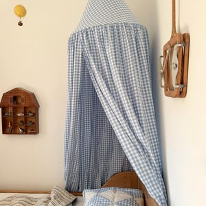 May include: A blue and white gingham fabric canopy suspended from the ceiling, featuring a conical top and long, flowing drapes that extend downwards to cover the headboard area of a wooden bed frame with a quilted blue and white patterned bedding.