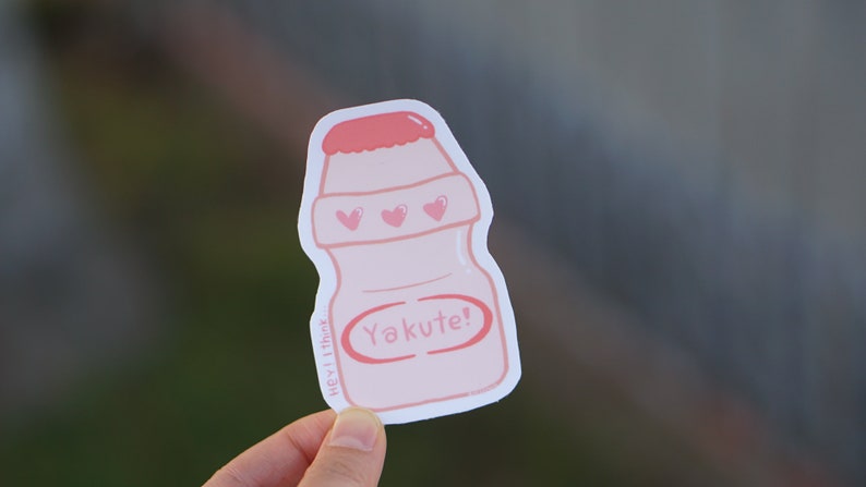 May include: Pink and white sticker of a Yakult bottle with a pink lid and three pink hearts on the front. The sticker says "Yakute!" in a pink circle.