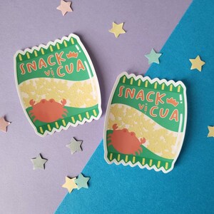 Puede incluir: Dos bolsas de aperitivos verdes y amarillas con un cangrejo rojo y el texto "Snack vi Cua" sobre un fondo morado y azul.