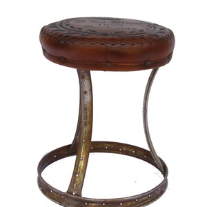 Puede incluir: Un taburete de cuero marr&oacute;n con un asiento circular y una base de metal. El asiento tiene un dise&ntilde;o decorativo y el texto "X.O. Special Reserve 1927 Bordeaux" impreso en &eacute;l.