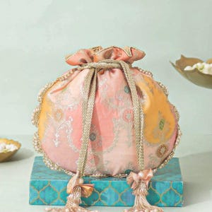 Puede incluir: Un bolso tipo saco con cordón de tela en tonos melocotón y amarillo con un patrón floral plateado metálico. El bolso está adornado con pequeñas perlas blancas en los bordes e incluye dos cordones dorados trenzados con borlas de cuentas.