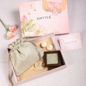 Puede incluir: Un set de caja de regalo con una tapa rosa con el texto AMYRA, una bolsa dorada con cordón y borlas, dos velas blancas pequeñas, un marco de madera cuadrado con un inserto verde estampado y una tarjeta con el texto Happy Diwali.