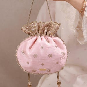 Pode incluir: Uma bolsa tipo saco rosa claro com bordados metálicos dourados e motivos florais de lantejoulas. A bolsa tem um topo franzido, uma alça trançada dourada e duas borlas douradas pendentes com contas. O tecido apresenta um brilho subtil.