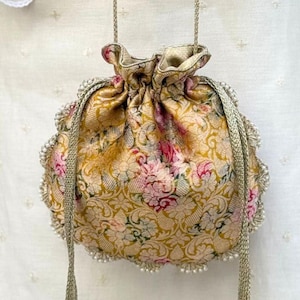 May include: A gold-toned drawstring pouch featuring a floral brocade pattern in shades of pink and green. The bag is accented with a scalloped edge of small white beads and includes two long, braided gold cords with beaded tassels at the ends.