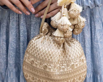 May include: A cream-coloured drawstring pouch bag featuring intricate gold embroidery and sequin detailing. The bag is adorned with a cluster of matching cream tassels and gold-toned decorative spheres, finished with a thin gold braided handle.