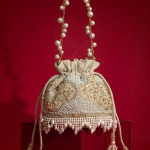 Op de afbeelding: Een beige buideltas met trekkoord, voorzien van ingewikkeld goudborduurwerk, witte parelversieringen en een bungelende parelfranje aan de onderkant. Het handvat is een lange gouden ketting met witte parels en de trekkoorden hebben parelkwastjes.