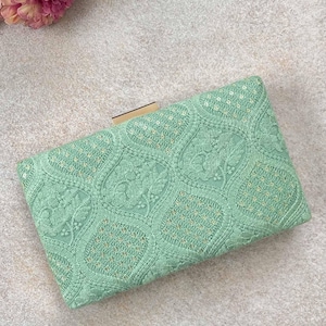 Puede incluir: Clutch rectangular verde menta con un intrincado patrón bordado. El clutch presenta un cierre dorado y está decorado con bordados florales. Flores rosas y melocotón en la esquina superior izquierda.