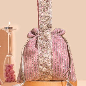 Peut inclure: Un sac bourse rose à cordon de serrage orné de rangées verticales de paillettes dorées et d'un panneau central avec une broderie florale dorée complexe. Le sac comprend une poignée assortie brodée et deux pompons dorés aux extrémités.