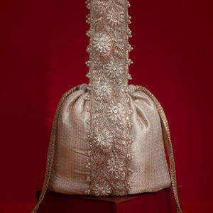May include: An ivory-colored drawstring bag with intricate floral embroidery and pearl accents. The bag features a long, embellished handle and a tassel, set against a red background. The bag is likely used for special occasions.