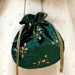 May include: A dark green velvet drawstring pouch bag featuring gold sequin floral embroidery. The bag has a braided gold cord handle and two long gold tassels with beaded accents at the base, creating a structured yet soft silhouette for carrying.