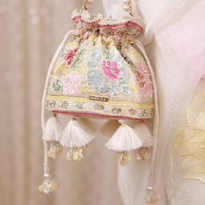 May include: A cream-colored drawstring pouch bag featuring intricate floral embroidery, sequins, and beadwork. The bag includes a gold-toned metal plate with the text AMYRA, a pearl-accented chain handle, and multiple white fabric tassels at the base.