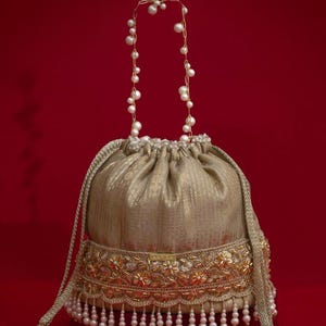 Könnte beinhalten: Ein goldfarbener Beutel mit Kordelzug, bestehend aus einem metallisch glänzenden Stoff, einem verzierten Stickband mit Blumenmotiven und einem unteren Saum aus weißen Perlen. Der Griff ist eine feine Goldkette mit weißen Perlen.