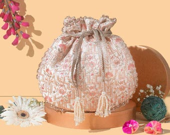 Puede incluir: Un bolso tipo saco de color crema con cordón, decorado con intrincados bordados florales en tonos rosa y naranja con patrones geométricos. El bolso se cierra con cordones plateados metálicos terminados en borlas de cuentas blancas.