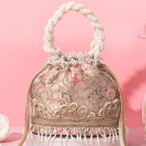 Könnte beinhalten: Eine Beuteltasche aus beigem Brokatstoff mit Blumenmuster und rosa Rosenakzenten. Die Tasche verfügt über einen gedrehten weißen Perlenhenkel, eine goldene Spitzenborte am Boden und einen Saum aus hängenden weißen Perlen am unteren Rand.