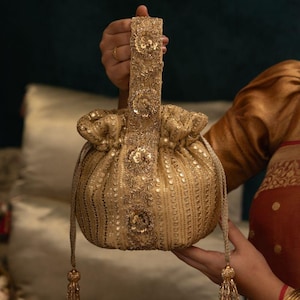 May include: A gold drawstring handbag featuring intricate embroidery, sequins, and metallic thread work. The bag has a single top handle and two side tassels with beaded accents, showcasing a dense pattern of reflective embellishments throughout.