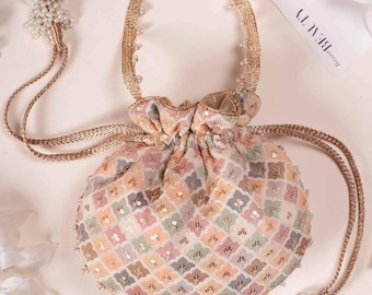 Puede incluir: Un bolso tipo saco con cordón y un patrón geométrico multicolor en tonos rosa, azul y dorado. El bolso está adornado con pequeñas lentejuelas y un borde metálico dorado. Incluye un asa trenzada dorada y dos cordones largos con borlas de cuentas blancas.