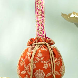 Könnte beinhalten: Ein orangefarbener Beutel mit Kordelzug und goldenem Blumenmuster, einem aufrechten Griff mit rosa-goldener Stickerei sowie zwei goldenen Kordeln, die in Büscheln aus kleinen, runden, metallisch goldenen Zierperlen enden.
