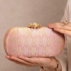 May include: A rectangular clutch bag in a soft pink hue, featuring an intricate woven pattern of stylized floral motifs in gold, silver, and yellow threads. The bag is finished with a metallic gold frame and a decorative gold flower-shaped clasp.