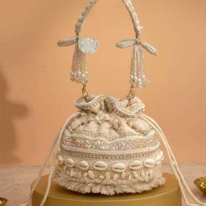 May include: Cream drawstring bag with detailed beadwork, seashells, and a beaded handle. The bag has a textured design, a fringed base, and decorative tassels. The bag is displayed on a gold-coloured stand.