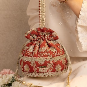 May include: A rust-colored potli bag featuring intricate gold floral embroidery, a drawstring closure with gold tassels, and a beaded handle. The bag has a rectangular gold label with the text AMYRA centered on the front, surrounded by decorative lace trim.