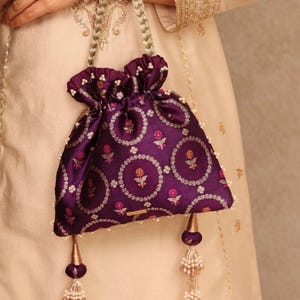 May include: A deep purple drawstring pouch bag featuring a repeating circular floral pattern in white and pink. The bag has a gathered top, braided handles, and two decorative hanging tassels with metallic gold cones and clear beaded accents.