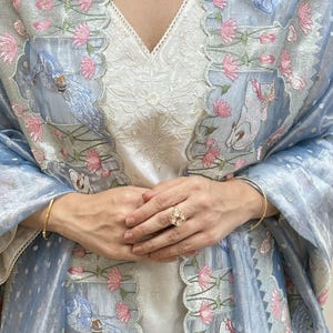 May include: A white embroidered tunic with a V-neck is paired with a light blue shawl featuring floral and bird embroidery. A gold ring and bracelet are visible. A beaded necklace adds a touch of elegance.