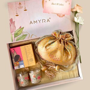 May include: A pink gift box with the word "AMYRA" printed on the lid. The box contains a gold brocade pouch, two small candle holders with floral embroidery, and a pink and white box with the word "PHOOL" printed on it. The box also includes a card that says "Best Wishes".
