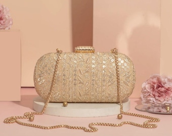 May include: Gold-colored, oval-shaped clutch with a beaded chain strap. The clutch is covered in intricate embroidery and sequins, with a decorative clasp. The background is a soft peach color, with pink flowers.