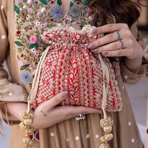 Może przedstawiać: Czerwona, haftowana torebka z ozdobnymi złotymi frędzlami. Torebka jest trzymana w dłoni i ma kwiatowy wzór.