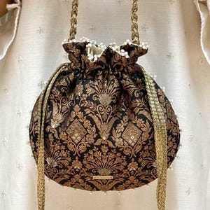 May include: A black and gold drawstring pouch bag featuring an intricate floral and geometric brocade pattern. The bag has a gathered top edge accented with small white beads, gold braided handles, and two long gold tassels with white bead detailing.