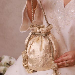 May include: A cream-coloured, drawstring pouch bag with intricate floral embroidery. The bag features a braided handle and decorative tassels with pearls. The bag is held by a person wearing a white embroidered outfit.