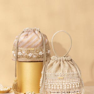 Può includere: Due pochette con coulisse in tessuto beige e dorato. La pochette a sinistra presenta un motivo a righe e ricami floreali. La pochette a destra è decorata con file di perle artificiali bianche e un manico intrecciato con nappe dorate.