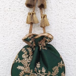 Könnte beinhalten: Ein dunkelgrüner Beutel mit Kordelzug und aufwendiger goldener Blumenstickerei. Die Tasche wird oben mit goldenen Kordeln zusammengezogen, die jeweils in einer dekorativen Quaste mit Perlenakzenten und Metalldrahtdetails enden.