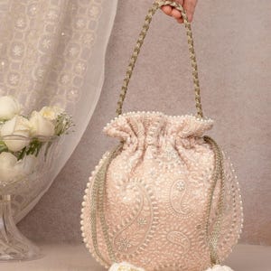 Pode incluir: Uma bolsa tipo saco de cor bege claro com bordados intrincados de flores e paisley em branco, acentuada com pequenas pérolas brancas ao longo das bordas e do cordão. A bolsa inclui uma alça de cordão dourado trançado e dois pingentes com pérolas.