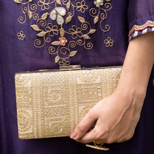 Op de afbeelding: Een gouden rechthoekige clutch met een decoratief patroon wordt vastgehouden tegen een donkerpaars kledingstuk. Het kledingstuk heeft bloemenborduurwerk met gouddraad en pastelaccenten. De clutch heeft een sluiting met een sluiting.