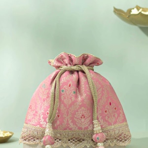 Puede incluir: Bolsa rosa con cordón y estampado floral dorado y ribete dorado. La bolsa tiene un cierre de cordón con cuerda dorada y borlas rosas. La bolsa está sobre una superficie blanca.