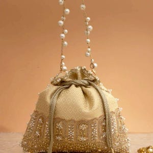 May include: A gold-toned drawstring pouch bag featuring a geometric pattern on the upper section and intricate embroidery with white beads and sequins on the lower border. The bag has a pearl-embellished chain handle and matching tassel drawstrings.