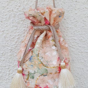 Puede incluir: Un bolso tipo saco con cordón cubierto de lentejuelas rosa pálido y patrones de tela floral. El bolso cuenta con un cierre de cordón dorado metálico con dos borlas largas de color crema a los lados y un asa de cordón a juego.