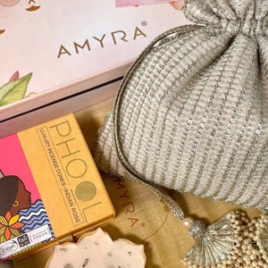 May include: A gift box set containing a white and gold decorative pouch, a candle, and a box of incense cones. The box is decorated with a floral design and the text "AMYRA". The pouch has a tassel with white beads. The candle is white with gold flecks. The incense cone box is yellow with a pink and black illustration and the text "PHOOL LUXURY INCENSE CONES - INDIAN ROSE". The card on top of the box says "Happy Diwali".