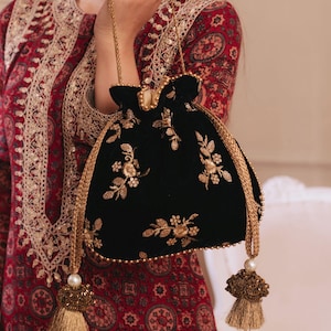 May include: A black velvet drawstring pouch bag featuring gold floral embroidery and sequins. The bag has a gold braided handle and two long decorative tassels at the base, each adorned with gold beads and multiple light brown fringe strands.