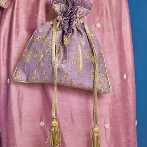 Puede incluir: Un bolso tipo saquito de color lavanda con bordados de hilo dorado en un patrón botánico. El bolso tiene un cierre fruncido con cordones trenzados dorados que terminan en largas borlas de cuentas doradas dispuestas en niveles.