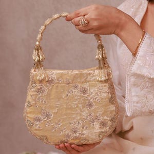 May include: A cream velvet handbag with floral embroidery and beaded detailing. The bag has a beaded handle and tassel embellishments. The design is ornate and elegant, suitable for special occasions.