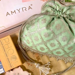 May include: A green and gold drawstring bag with a pearl tassel, a box with the text "AMYRA" and "Best Wishes", a box of incense cones, and a pink candle with gold flakes.
