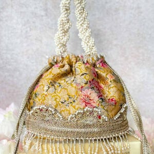 May include: A gold drawstring pouch bag featuring a floral pattern in shades of pink and yellow. The bag is adorned with a braided pearl handle, intricate metallic gold embroidery, and a decorative fringe of hanging white beads along the base.