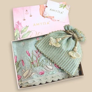 Puede incluir: Un juego de regalo con una bufanda bordada azul claro con cisnes y diseños florales, una bolsa a juego y una caja rosa con la marca "AMYRA". La bolsa tiene borlas y un cierre de cordón.