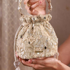 Könnte beinhalten: Eine goldene Beuteltasche mit aufwendigem Spitzenüberzug, eingebetteten Spiegelakzenten und einem perlenbesetzten Griff. Die Tasche verfügt über einen Kordelzug mit perlenbesetzten Quasten und ein kleines rechteckiges Metallschild mit der Aufschrift AMYRA.