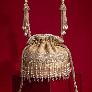 May include: A gold drawstring pouch bag featuring intricate embroidery, a top handle adorned with hanging beaded tassels, and a base fringe of dangling white pearls and clear crystals. The bag has a gathered top closure with long braided gold cords.