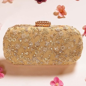 Op de afbeelding: Een goudkleurige, ovale clutch met ingewikkelde bloemenborduurwerk en pailletten. De clutch heeft een rechthoekige sluiting en is omgeven door verspreide roze en paarse bloemen.